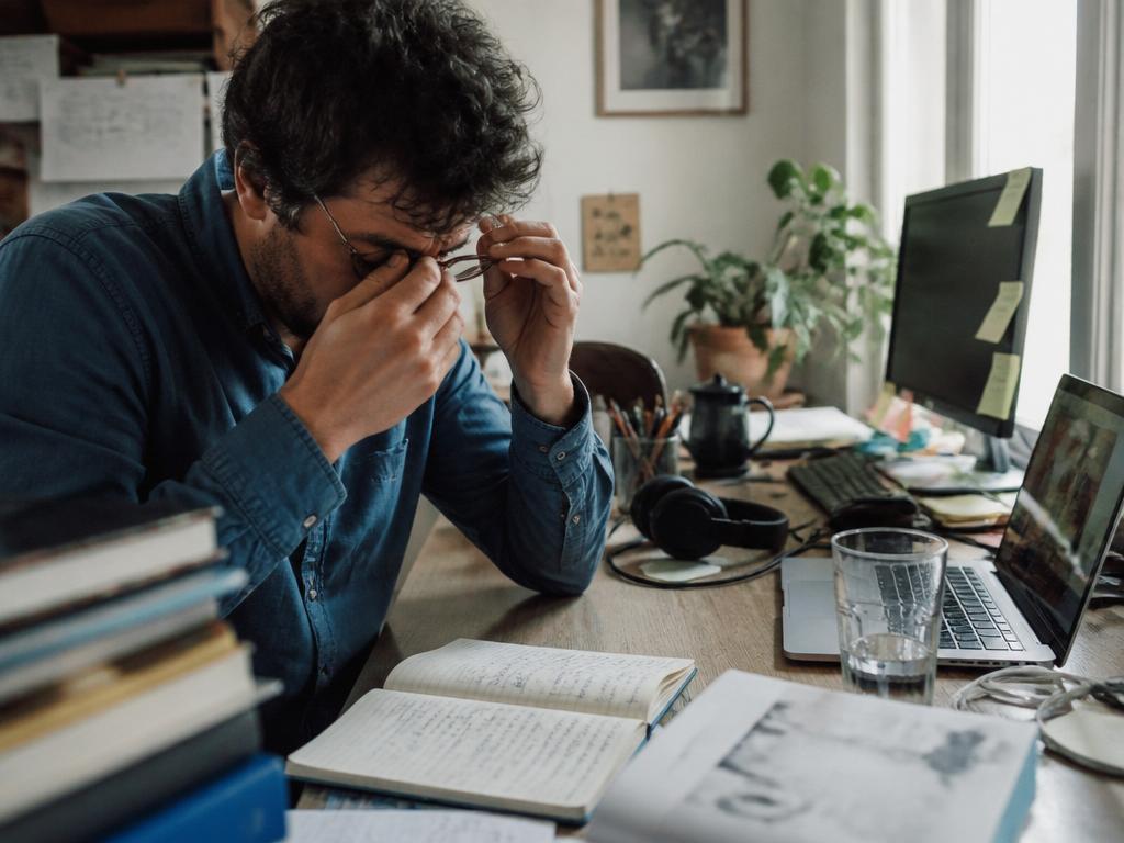A man at a cluttered home desk rubs his eyes and pushes up his glasses, gathering himself to refocus, in bright window light.
