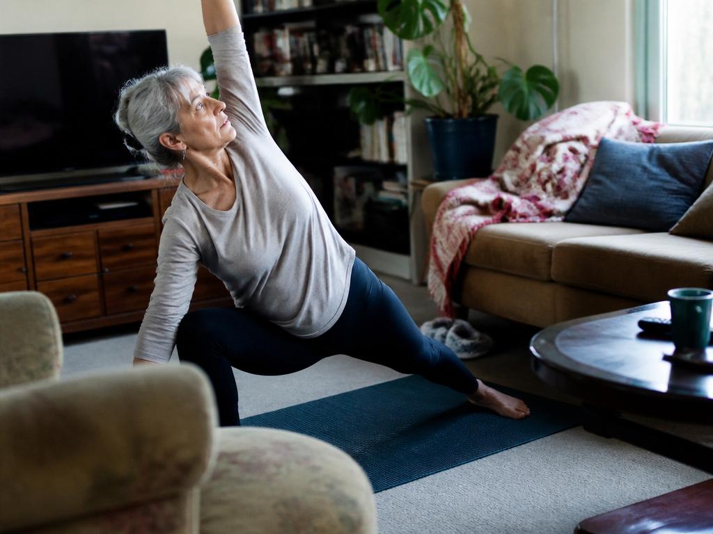 A woman in her late sixties holds a deep lunge stretch with one arm reaching up on a yoga mat in a bright, cluttered living room.