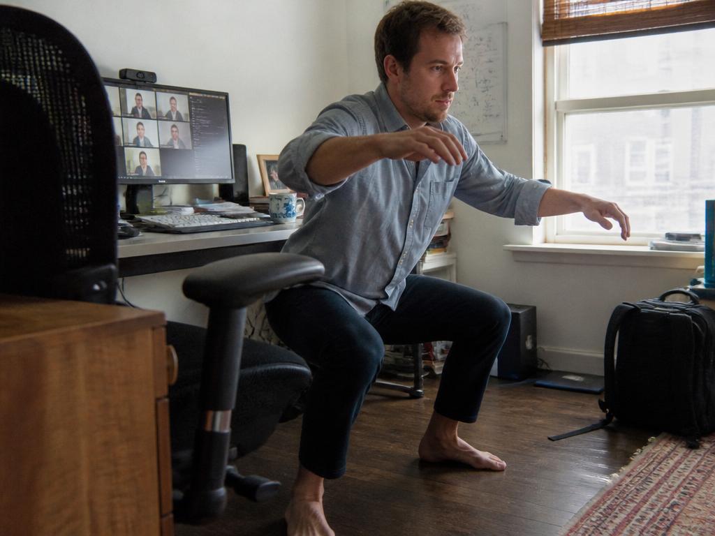 A man does bodyweight squats beside his desk chair during a quick work break in a bright, cluttered home office.